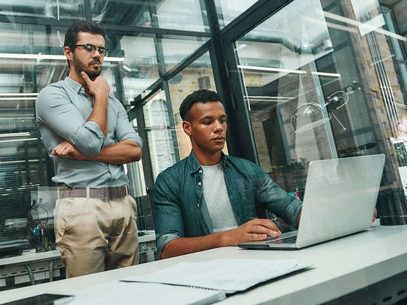 Two men in casual wear looking at screen of laptop and thinking about new project while working in modern office